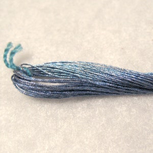 Puede incluir: Primer plano de un ovillo de hilo de bordar azul. El hilo tiene un brillo metálico, con tonos de azul claro y oscuro. El extremo del hilo está en bucle, mostrando los hilos individuales. Ideal para proyectos de bordado y manualidades.