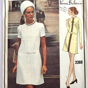 Pode incluir: Uma página de revista de moda vintage com uma mulher em um vestido branco, chapéu e joias. O vestido tem mangas curtas e cinto. Um desenho de um vestido com o número 2360 está à direita. O logotipo Pierre Balmain está no topo.