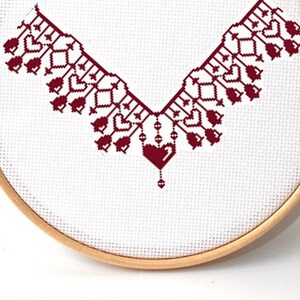 May include: A white embroidery hoop with a red cross-stitch design. The design features a V-shaped pattern with a heart at the bottom.