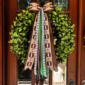May include: A circular green boxwood wreath adorned with a large burlap bow featuring a brown and white football pattern. Multiple long, hanging ribbons in various shades of green, brown, and white with football and polka dot prints hang below.