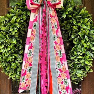 May include: A decorative wreath featuring a dense circle of green leaves adorned with a large, multi-layered bow. The bow consists of ribbons with pink and white checkered patterns, pineapple prints, green gingham, and a central strand of pink tassels.