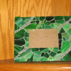 May include: A green glass mosaic picture frame with silver accents. The frame is made of small pieces of glass that are arranged in a pattern. The frame is sitting on a wooden surface.