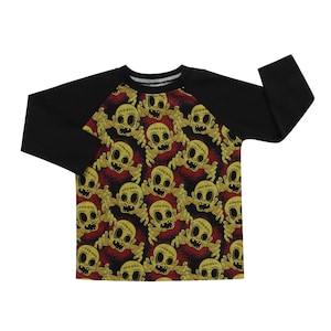 Puede incluir: Camiseta de manga larga negra y amarilla con diseño raglan. La camiseta presenta un patrón repetido de cabezas de zombis de dibujos animados con caras cosidas y manos extendidas.