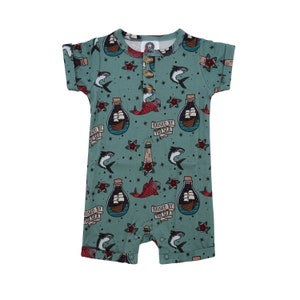 May include: A teal blue baby onesie with a nautical theme. The onesie has a repeated pattern of a bottle with a ship inside, a red fish, a lighthouse, and a shark. The text "Raised by the Sea" is repeated on the onesie.