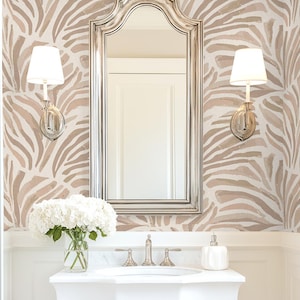 May include: A bright, white bathroom with a patterned wallpaper in shades of beige and white. A silver-framed mirror hangs above a white sink with a bouquet of white flowers. Two sconces flank the mirror, and the floor has a patterned tile.