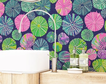 Puede incluir: Un baño con un papel tapiz estampado en verde azulado, verde y rosa con un diseño repetitivo de erizos de mar. Un espejo redondo cuelga sobre un lavabo de porcelana blanca en un tocador de madera.