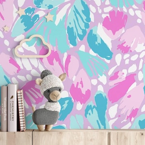 May include: A crochet llama toy with a grey body and white head stands beside three books. The background features wallpaper with a repeating abstract pattern of pink, light blue, and white shapes, accented by small wooden star and cloud cutouts.