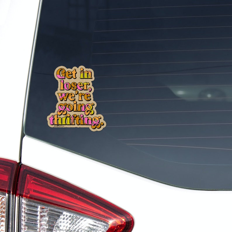 Op de afbeelding: Een auto sticker voor het achterraam met de tekst "Get in loser, we're going thrifting." in een retro lettertype met roze, oranje en bruine kleuren.