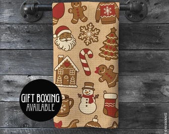 May include: A beige Christmas-themed tea towel featuring festive illustrations such as gingerbread men, Santa Claus, and a snowman. The towel hangs on a black metal bar against a wooden backdrop. The text "GIFT BOXING AVAILABLE" is in a black circle.