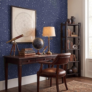 May include: A dark wood desk and matching leather chair set against a navy blue wall with a white star pattern. The desk holds a brass telescope, a celestial globe, a lamp with a beige shade, and stacked books. A tall bookshelf stands nearby.