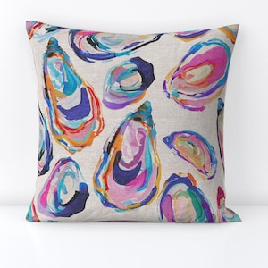 May include: A decorative pillow featuring a repeating pattern of colorful oyster shell illustrations. The shells are painted with vibrant hues of blue, pink, orange, and purple on a neutral background. The pillow is square and appears to be made of a linen-like fabric.