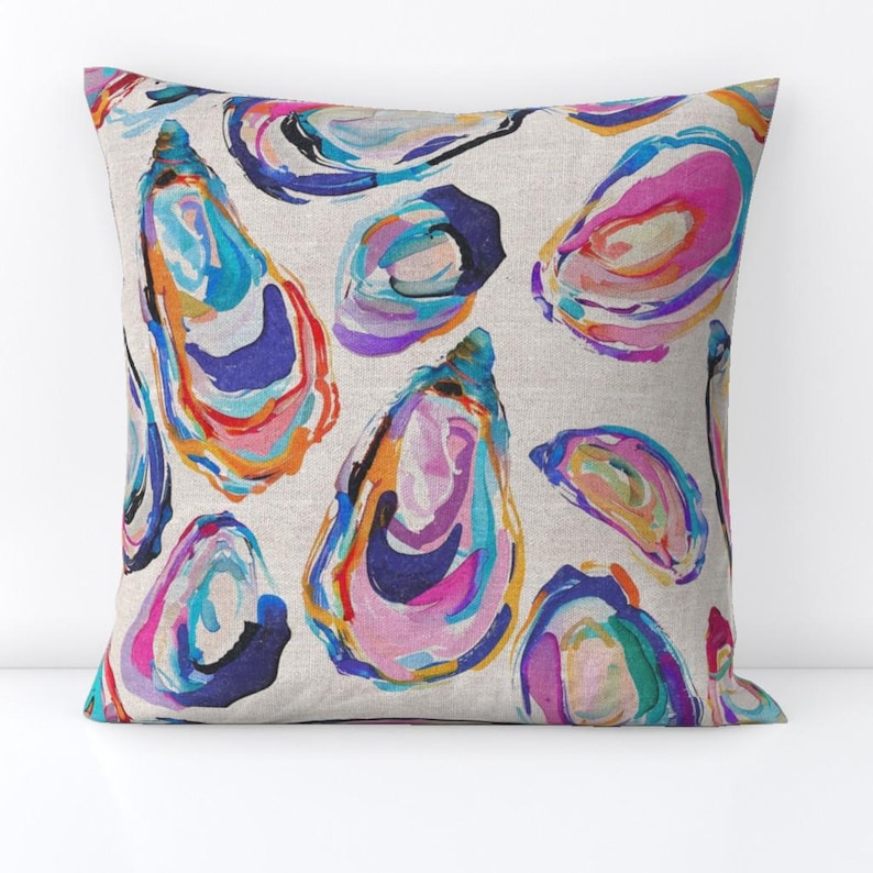 May include: A decorative pillow featuring a repeating pattern of colorful oyster shell illustrations. The shells are painted with vibrant hues of blue, pink, orange, and purple on a neutral background. The pillow is square and appears to be made of a linen-like fabric.