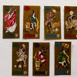 May include: A collection of seven postage stamps with a gold background. Each stamp features a stylized illustration of a person in traditional attire, some with drums. The stamps have text and various denominations.