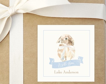 May include: A white gift box with a white satin ribbon tied in a bow. A square gift tag with a watercolor illustration of a brown bunny wearing a blue ribbon is attached to the box. The tag reads "It's a Boy" and "Luke Anderson".