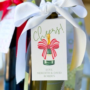 May include: A bottle of wine with a white ribbon and a tag that says "Cheers!" in green script. The tag features a watercolor illustration of a champagne bottle with a red and white bow. The tag also includes the text "Love, Meredith & David Roberts."