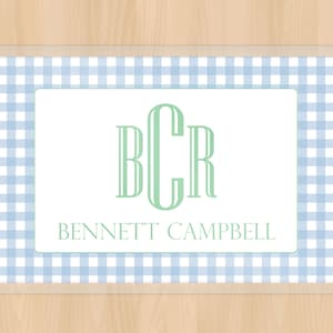 May include: A personalized rectangular placemat with a light blue and white gingham pattern border. The center features a white rectangle with the initials "BCR" in green and the name "BENNETT CAMPBELL" below.