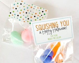 May include: Two clear cellophane bags with colorful Halloween-themed toys. One bag contains a green and pink toy, and the other has blue and orange toys. Each bag has a decorative tag that says "Squishing You a Happy Halloween!"