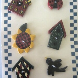 May include: A collection of handcrafted decorative buttons in various shapes, including birdhouses, a ladybug, a sunflower, and a bird. The birdhouse buttons feature red, green, and black designs. The ladybug is red with black spots. The word "Handcrafted" is printed at the bottom.