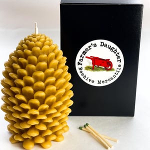 May include: A yellow beeswax candle shaped like a pine cone with a white wick. The candle is packaged in a black box with a white sticker that says "Farmer's Daughter Beeshive Mercantile" and depicts a red wheelbarrow.
