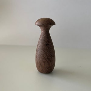 Könnte beinhalten: Ein dunkelbrauner, holzerner Flaschenverschluss in Form eines Pilzes mit einer glatten, abgerundeten Oberseite.