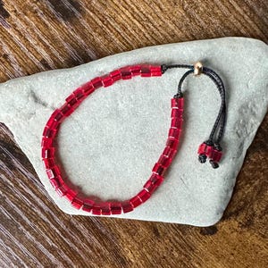 May include: A red beaded bracelet with a black adjustable cord and gold-coloured bead. The bracelet is composed of small, rectangular red beads. The cord is tied with two small red beads at the ends. The bracelet is displayed on a grey stone.