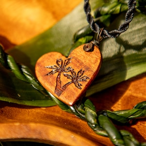 May include: A heart-shaped wooden pendant with a palm tree design. The pendant is suspended from a black cord necklace. The pendant is brown with a dark brown palm tree design. The background is a mix of green and brown.