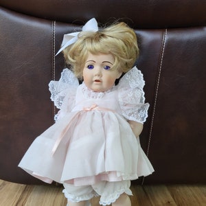 May include: A porcelain doll with blonde hair, wearing a white dress with lace trim and black shoes. The doll has blue eyes and a pink bow in her hair.