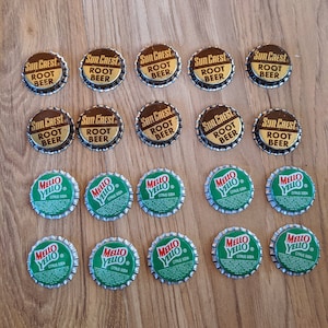 May include: A collection of 15 vintage bottle caps, 9 are gold with the text "Sun Crest Root Beer" and 6 are silver with the text "Mello Yello Citrus Soda".