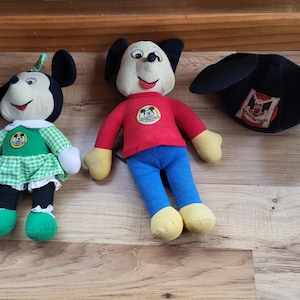 May include: Three plush Mickey Mouse toys. One is a girl Mickey Mouse wearing a green and white plaid dress with a red bow. Another is a boy Mickey Mouse wearing a red shirt and blue pants. The third is a black Mickey Mouse hat with a red and white logo.