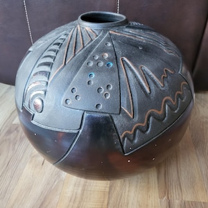 May include: A spherical black ceramic vase featuring a textured upper section with carved geometric patterns, including triangles, wavy lines, and circular indentations. The surface displays subtle copper-toned highlights and small blue accents.