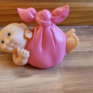 May include: A vintage baby doll figurine, featuring a smiling face with blue eyes and small hands. The doll is wrapped in a pink sack with a bow-like top, and its tiny feet are visible. The doll is lying on a wooden surface.