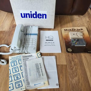 May include: A white Uniden HQ Series feature phone with a keypad, handset, and coiled cord, alongside its box and paperwork. The box has the Uniden logo. Additional items include a modular jack and a keypad overlay.