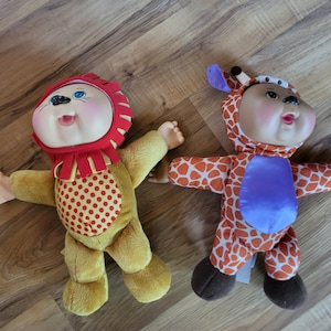 May include: Two Cabbage Patch Kids dolls dressed in animal costumes. One doll is wearing a red and yellow lion costume with brown fur. The other doll is wearing an orange and white giraffe costume with brown fur.