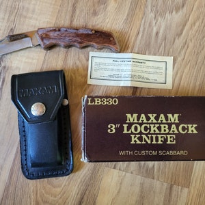 May include: A brown box with gold lettering reads "Maxam 3" Lockback Knife with Custom Scabbard". A brown handled folding knife with a black leather sheath is next to the box. The knife has a full lifetime warranty.