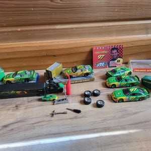 May include: A collection of toy race cars and accessories, including miniature green and yellow race cars with the number 97, a semi-truck, and a promotional card. The items are arranged on a wooden surface.