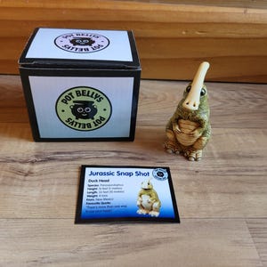 May include: A green and brown ceramic figurine of a duck-billed dinosaur with a long, thin beak. The figurine is standing on a wooden surface. The figurine is labeled "Jurassic Snap Shot" and "Duck Head".