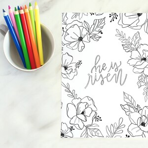 May include: A black and white printable colouring page featuring a floral design with the text "He is risen" in a handwritten font. The page is surrounded by a border of flowers and leaves.