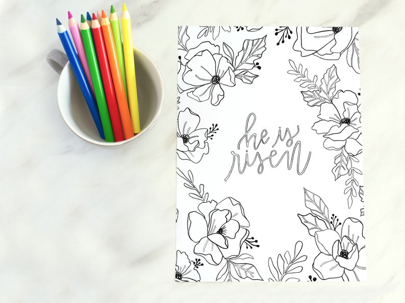 May include: A black and white printable colouring page featuring a floral design with the text "He is risen" in a handwritten font. The page is surrounded by a border of flowers and leaves.