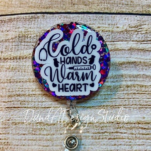 May include: A round, white, glitter-filled badge holder with a black and white design that reads "Cold Hands Warm Heart". The badge holder is attached to a clear plastic retractable badge reel.