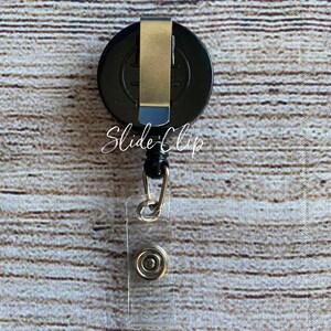 May include: Black retractable badge holder with a silver clip and a clear plastic strap. The badge holder has the text "Slide-Clip" printed on it.