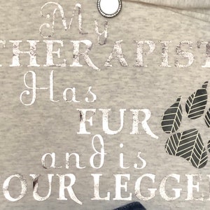 Puede incluir: Una sudadera gris con el texto "My therapist has fur and is four legged" en letras plateadas con purpurina. Una huella de pata negra se encuentra en la esquina inferior derecha.