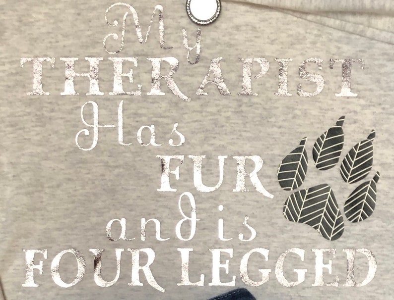 Puede incluir: Una sudadera gris con el texto "My therapist has fur and is four legged" en letras plateadas con purpurina. Una huella de pata negra se encuentra en la esquina inferior derecha.