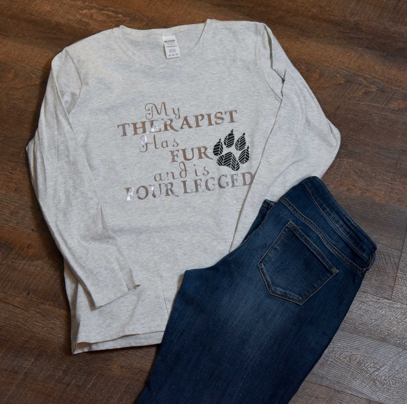 Puede incluir: Una camiseta de manga larga gris claro con el texto "My therapist has fur and is four legged" y un gr&aacute;fico de huella de pata.