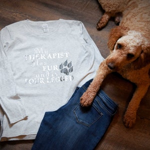Puede incluir: Una camiseta de manga larga gris claro con el texto "My therapist has fur and is four-legged" y un gr&aacute;fico de huella de pata. Un perro marr&oacute;n est&aacute; acostado en el suelo junto a la camiseta y un par de pantalones vaqueros azules.