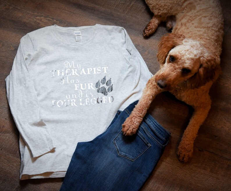 Puede incluir: Una camiseta de manga larga gris claro con el texto "My therapist has fur and is four-legged" y un gr&aacute;fico de huella de pata. Un perro marr&oacute;n est&aacute; acostado en el suelo junto a la camiseta y un par de pantalones vaqueros azules.