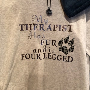 Puede incluir: Sudadera gris con el texto "Mi terapeuta tiene pelo y es de cuatro patas" y un gr&aacute;fico de una huella de pata.