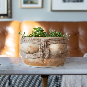 May include: A ceramic planter shaped like a bust with a rope tied around it. The planter is painted in a light brown color and has a rustic look. The planter is filled with a green succulent plant.