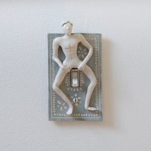May include: A ceramic light switch cover with a white figurine of a person in a seated position. The switch cover is light blue with a white patterned design.