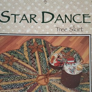 May include: A Star Dance tree skirt packaging featuring a circular fabric skirt with a patchwork design of green, red, and tan star patterns. The packaging includes the text STAR DANCE Tree Skirt and an image of the skirt with small toy decorations.