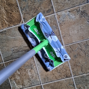 May include: A green Swiffer mop head attached to a gray handle, featuring a reusable blue fabric cover with a white and black butterfly pattern wrapped around the base. The cleaning tool is positioned on a brown and gray tiled floor surface.